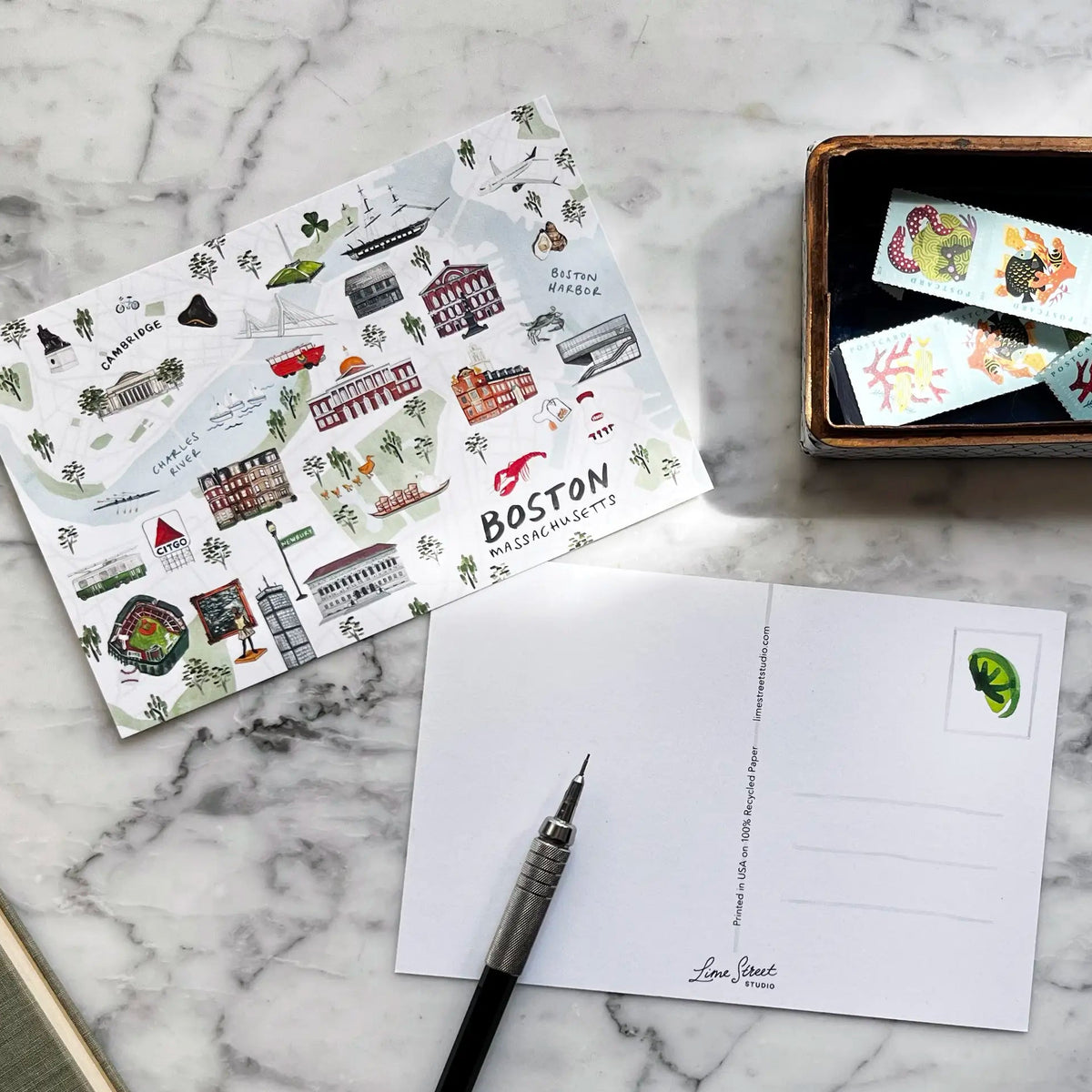 A styled flat-lay of a postcard set on a white marble surface, featuring a printed Boston postcard sheet, an open postcard with a green leaf stamp, a black pen, a small wooden box with colorful illustrated strips, and a dark blue mug.